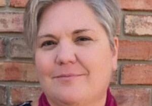 A woman with short hair is standing in front of a brick wall.